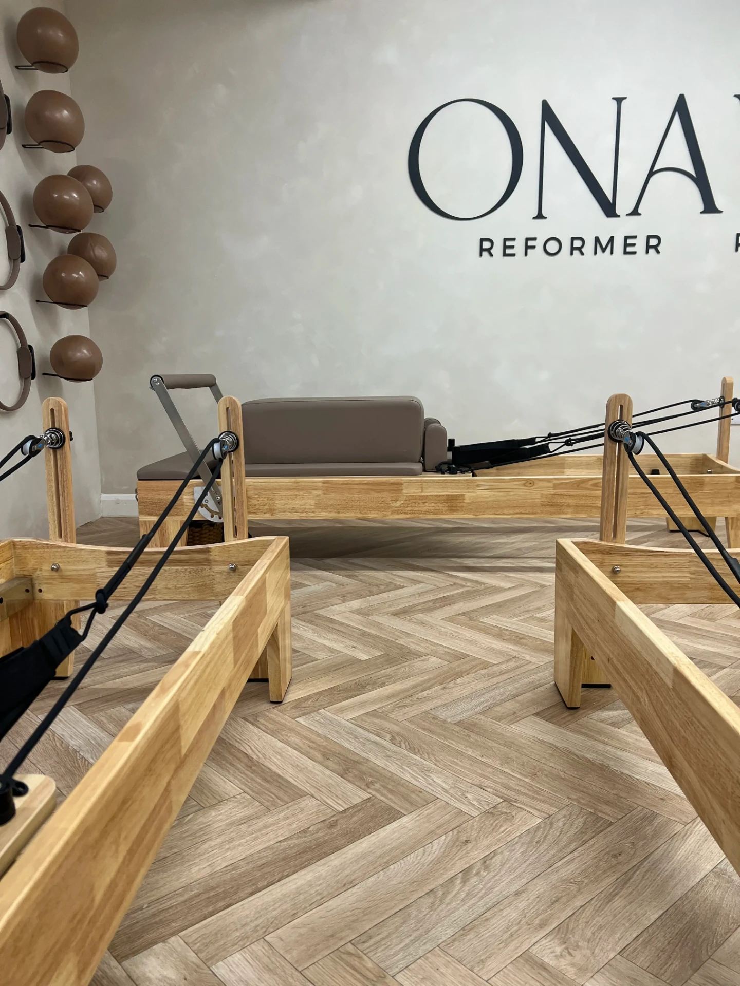 Row of wooden reformers on herringbone flooring beneath the Onavie Reformer Pilates wall signage
