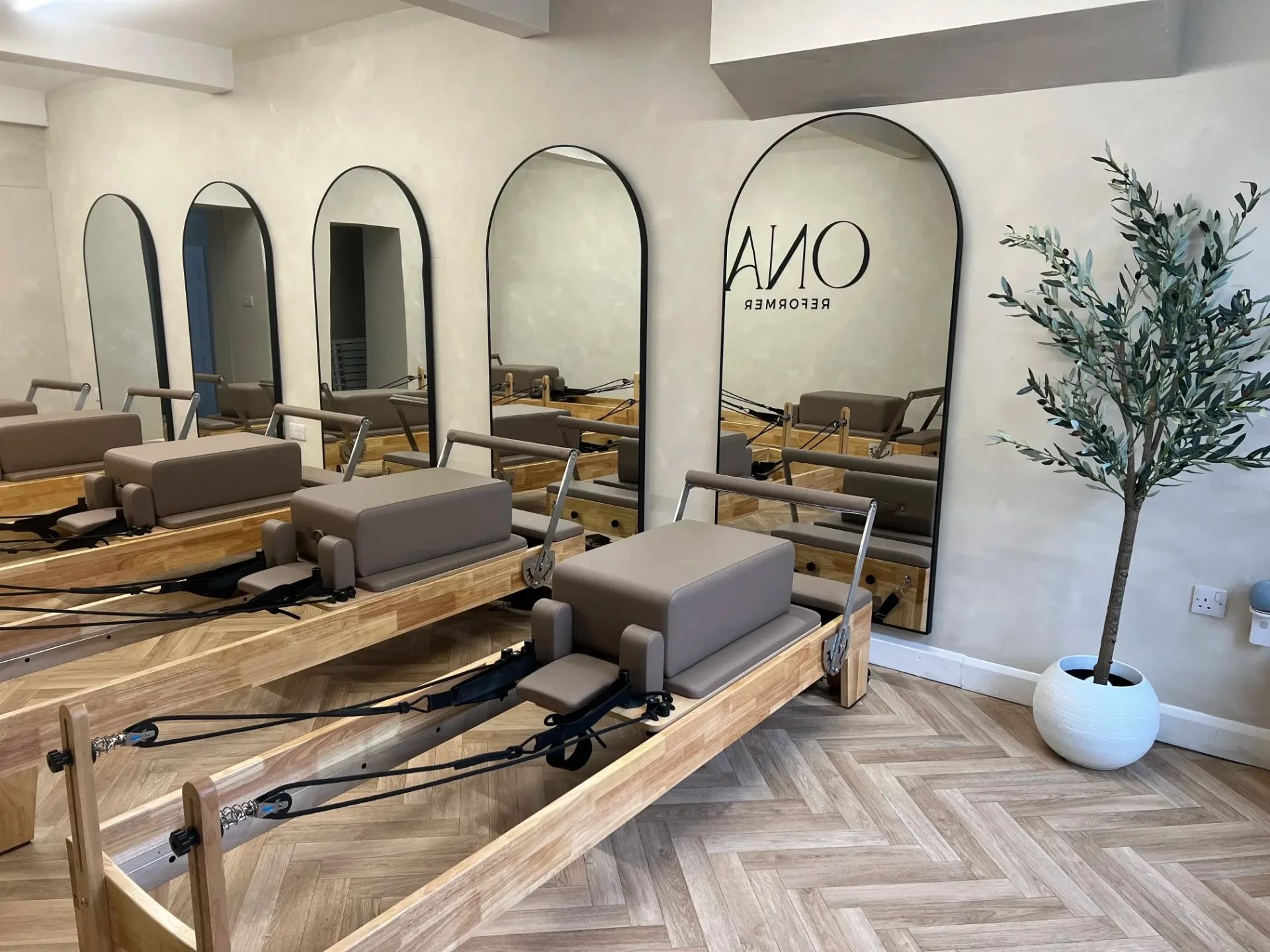 Arched mirrors along the Onavie studio wall, reformers on herringbone flooring beside an olive tree